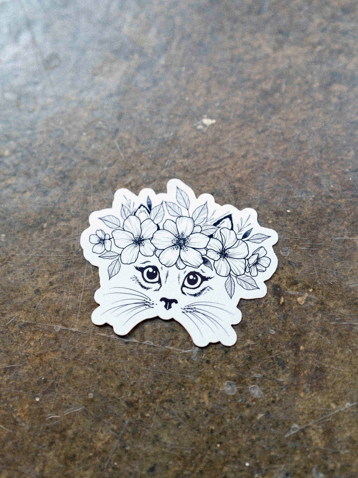 A black and white vinyl sticker of a cat with a floral crown illustration by Janice Danger on a concrete background.