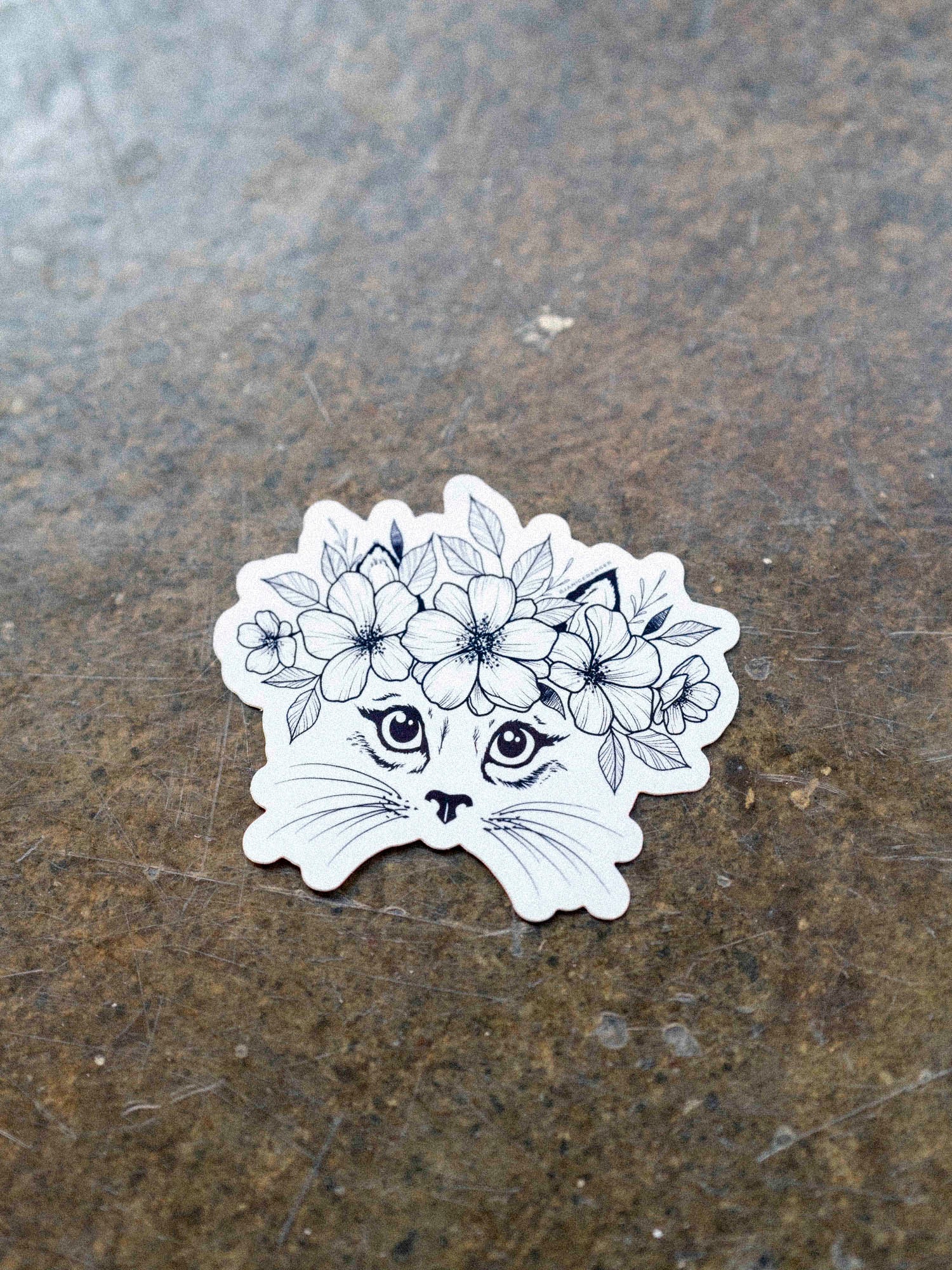 A black and white vinyl sticker of a cat with a floral crown illustration by Janice Danger on a concrete background.