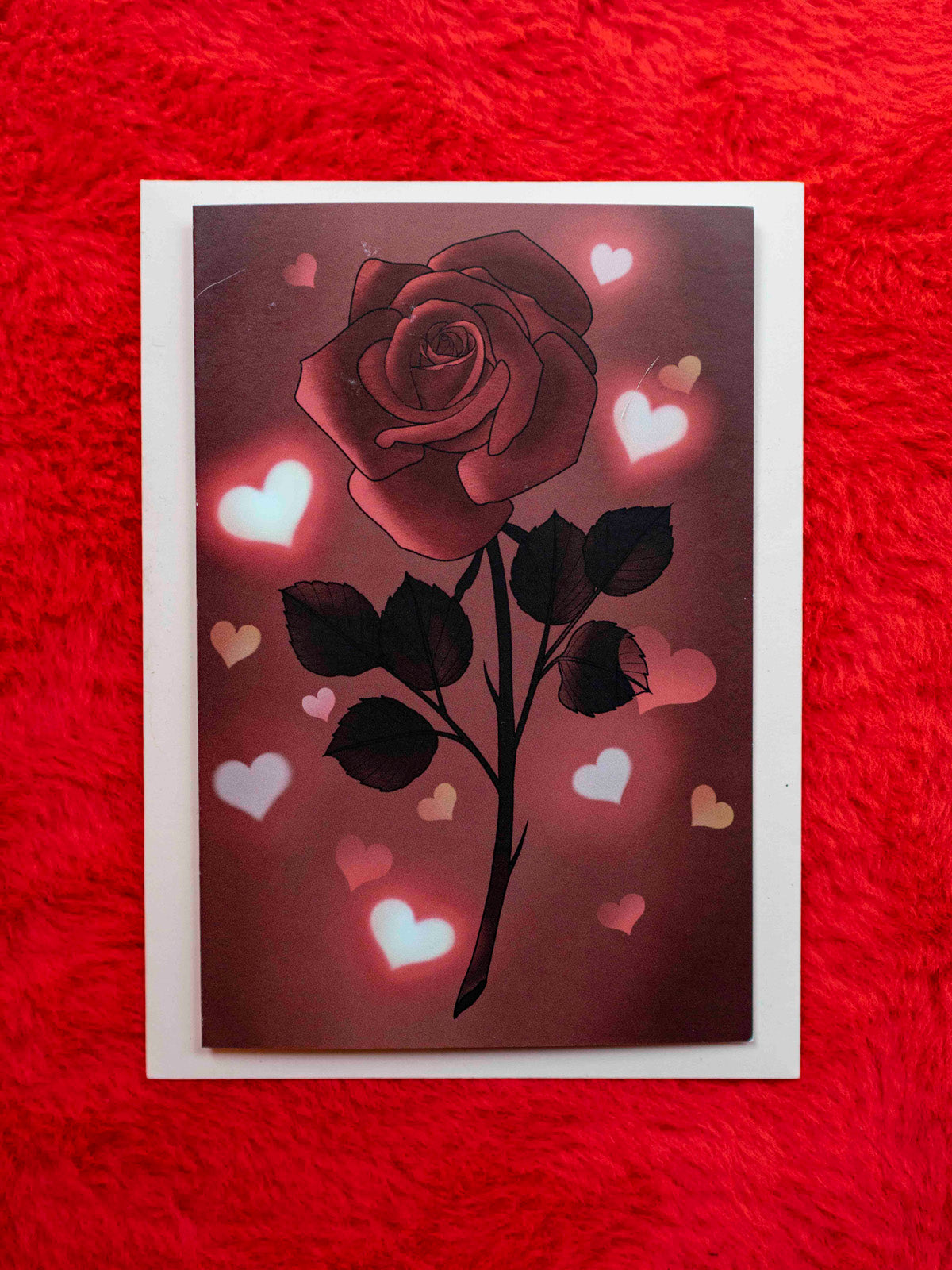 A folded greeting card with a red rose and hearts illustration on the front, with a pink envelope included.