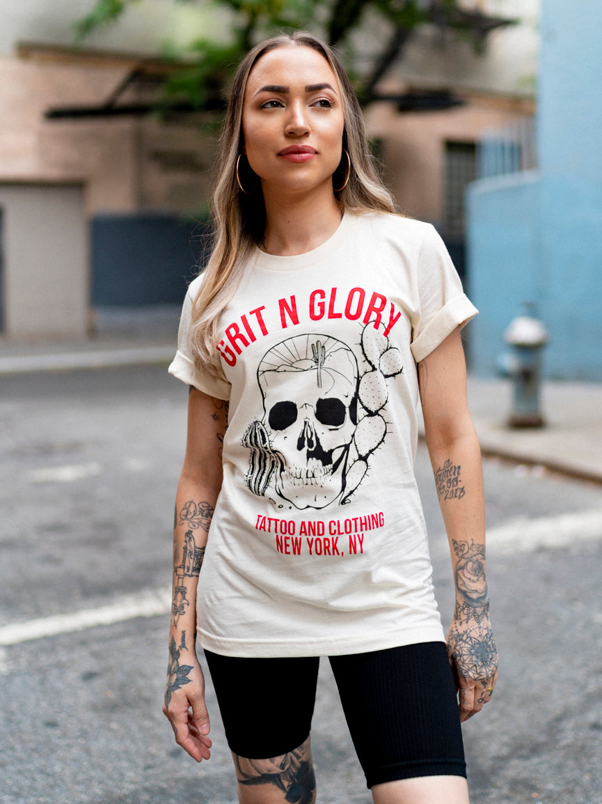 A person wearing a white crew neck t-shirt with a graphic design of a desert themed skull and Grit N Glory red text.