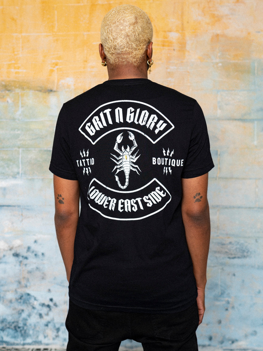 A person wearing a black, short-sleeved T-shirt with a scorpion graphic on the back and the text 'Grit N Glory'.