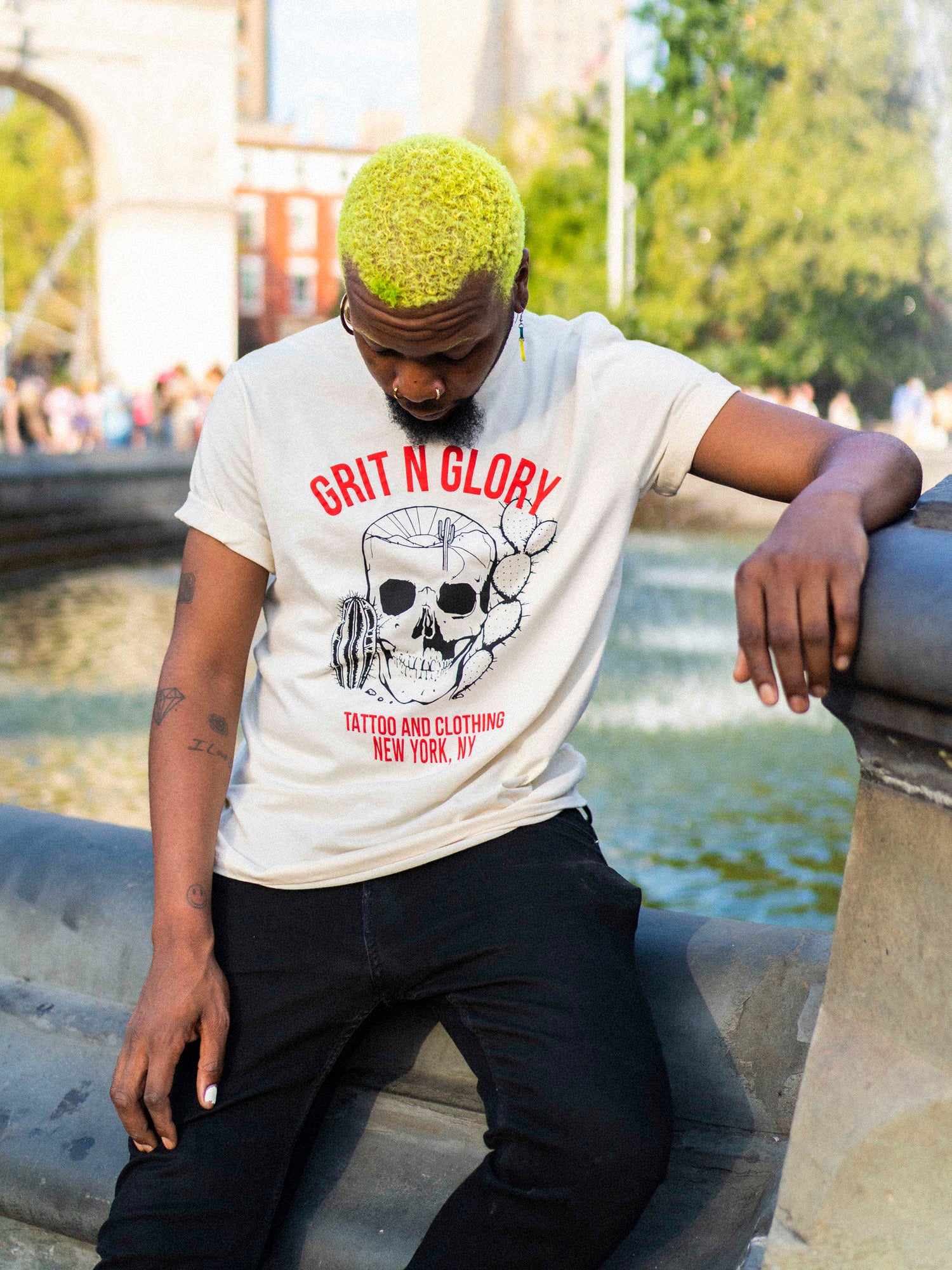 A person wearing a white crew neck t-shirt with a graphic design of a desert themed skull and Grit N Glory red text in front of the Washington Square Park Fountain.