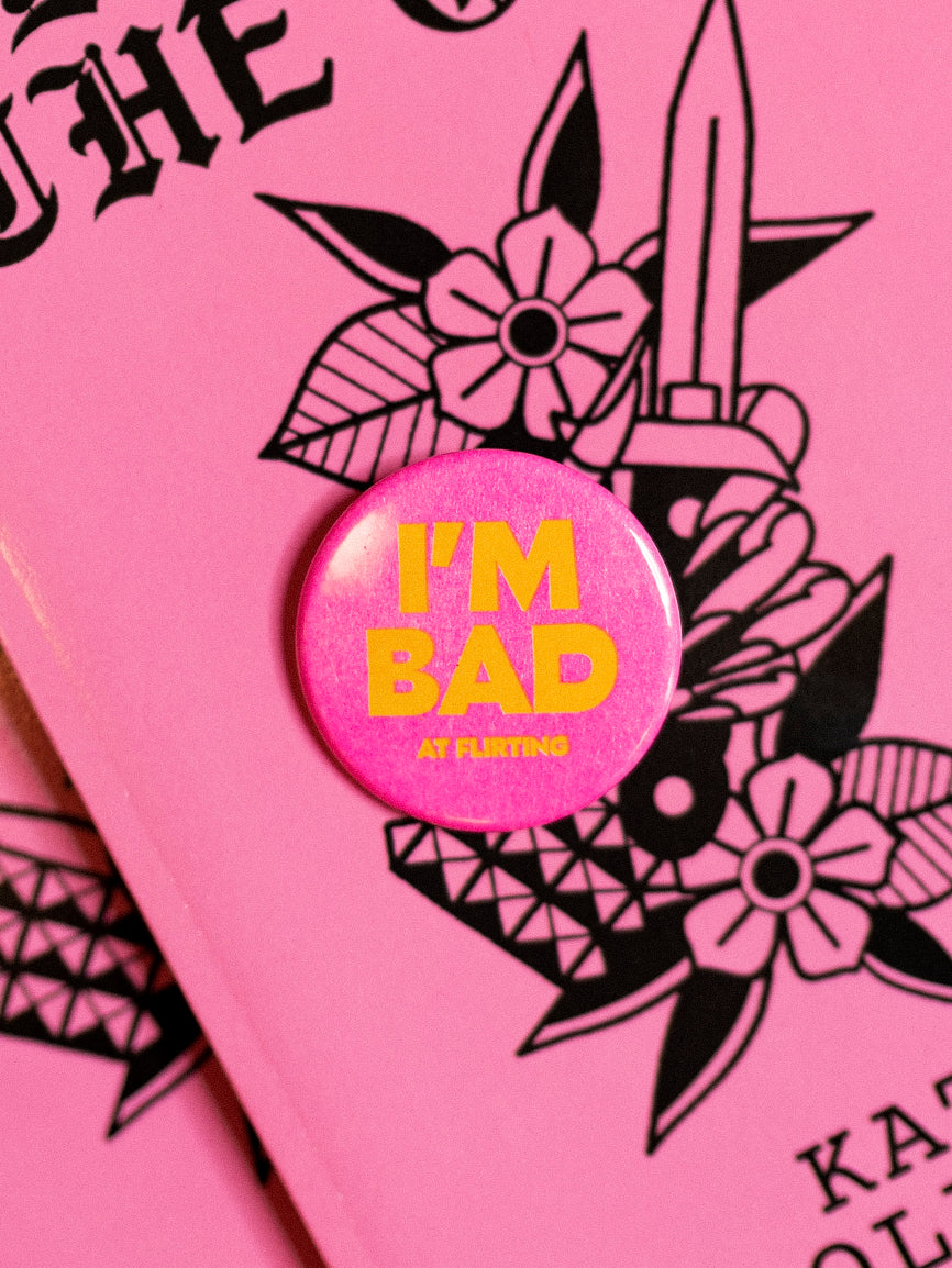 Classic round hot pink button reads "I'm Bad at flirting" in yellow against a black and pink traditional tattoo themed book. 