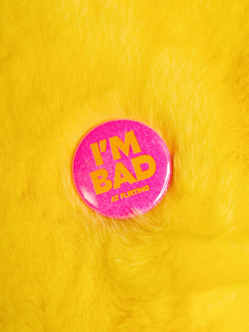 Classic round hot pink button reads "I'm Bad at flirting" in yellow against a fuzzy yellow background.