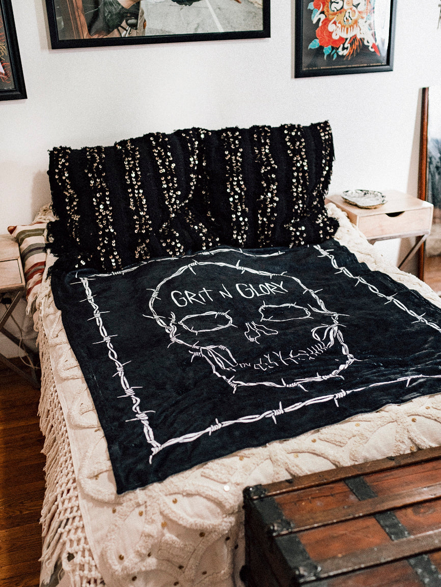 A black blanket with a white skull design and the text 'GRIT N GLORY' on a bed.