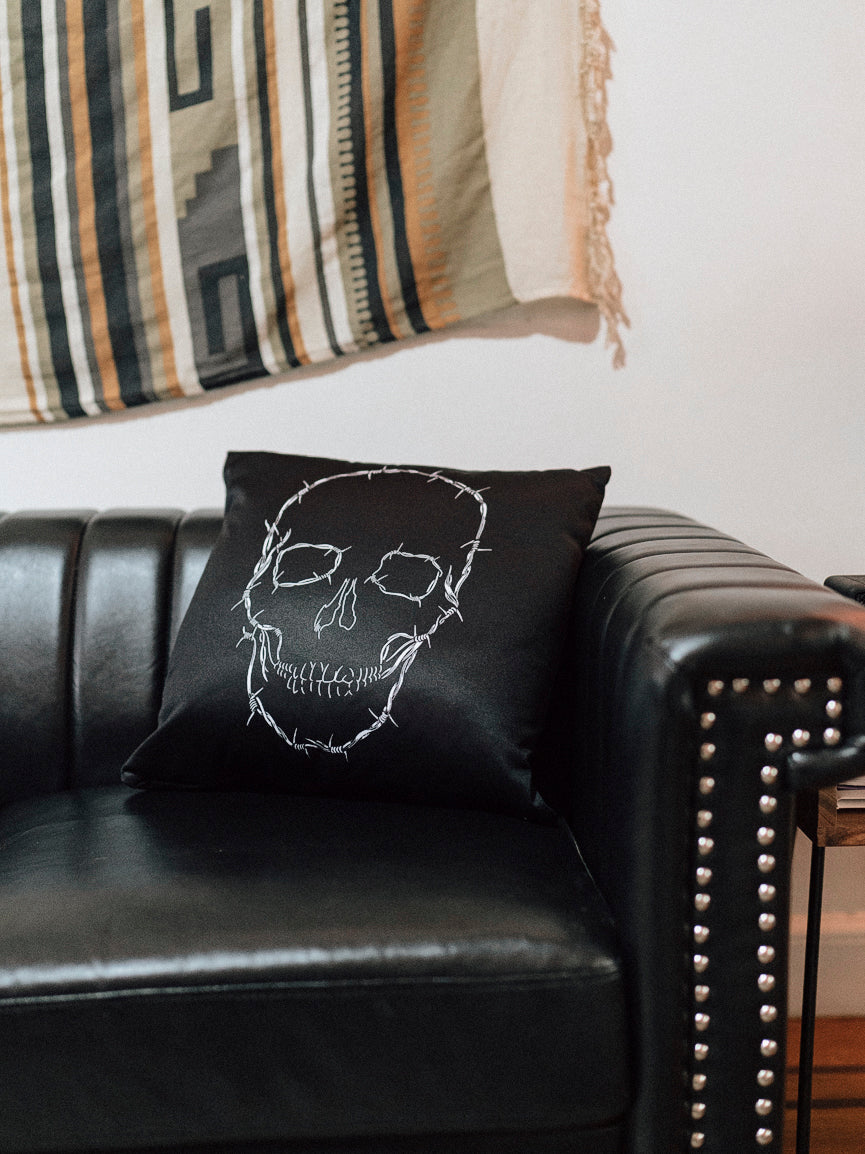A black throw pillow with a white skull design on it, placed on a leather sofa with a striped rug in the background.
