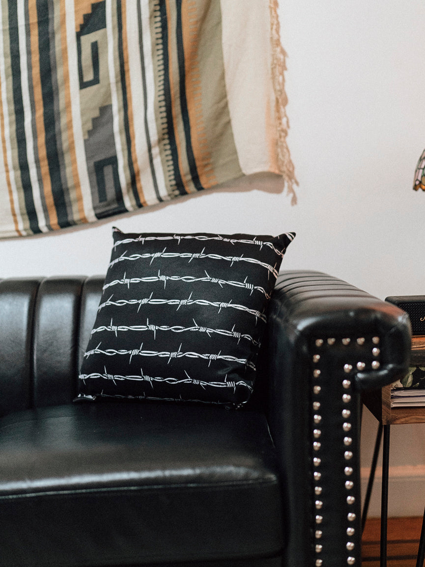 A black throw pillow with a white barbed wire pattern on it, placed on a leather sofa armrest with a patterned lampshade in the background.