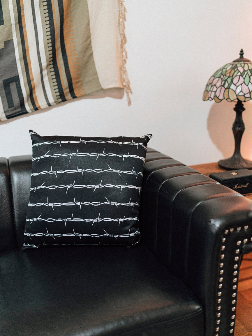 A black throw pillow with a white barbed wire pattern on it, placed on a leather sofa armrest with a patterned lampshade in the background.