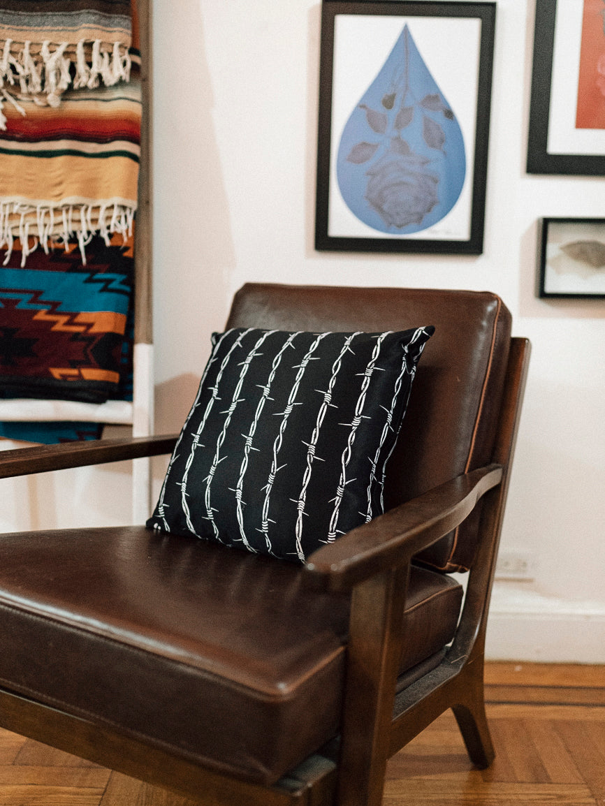 A black throw pillow with a white barbed wire pattern on it, placed on a leather chair.