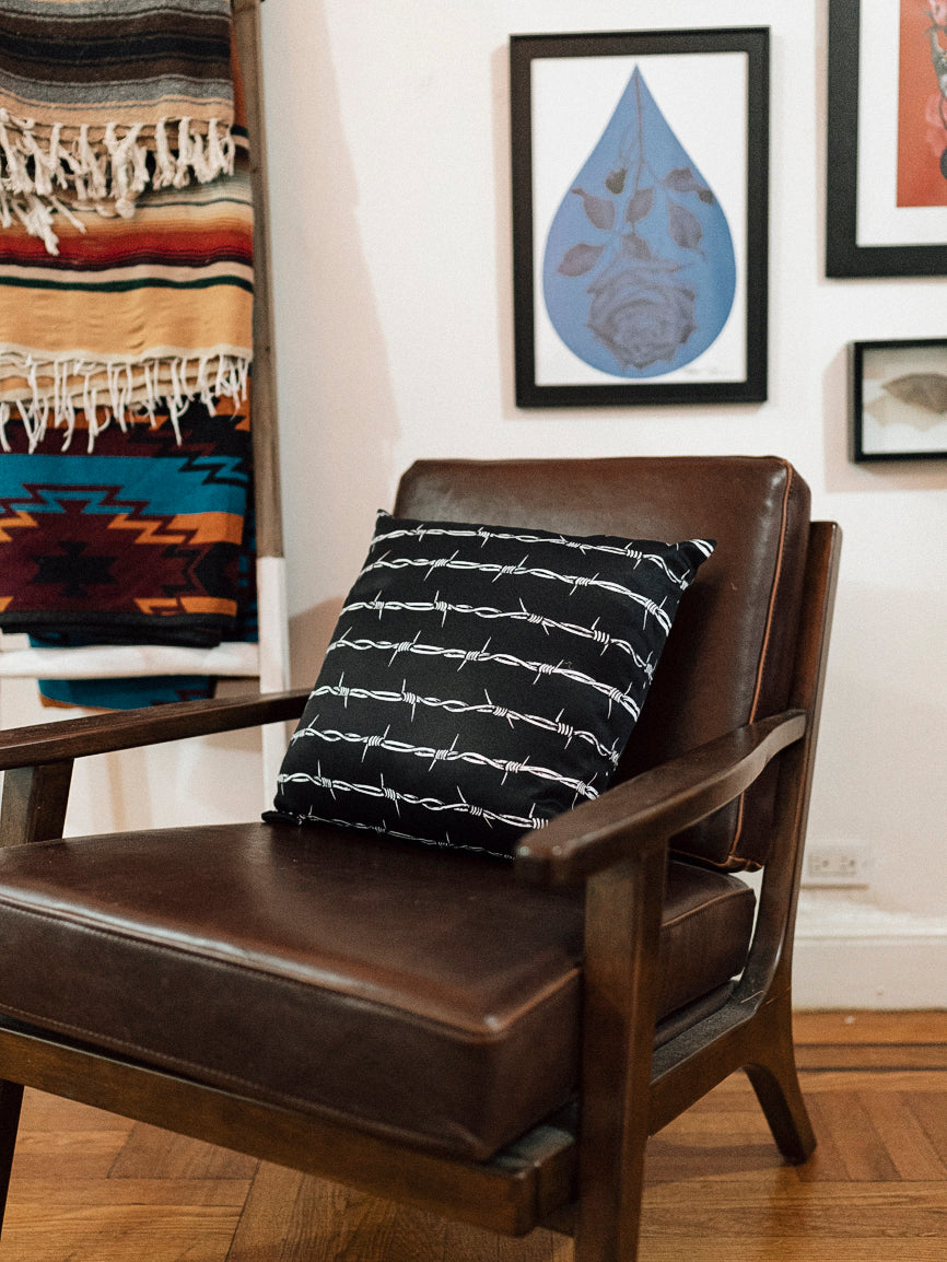 A black throw pillow with a white barbed wire pattern on it, placed on a leather chair.