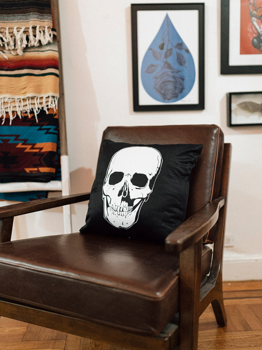 A black throw pillow with a white skull design on it, placed on a leather sofa with a striped rug in the background.