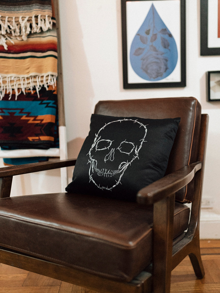 A black throw pillow with a white skull design on it, placed on a leather sofa with a striped rug in the background.