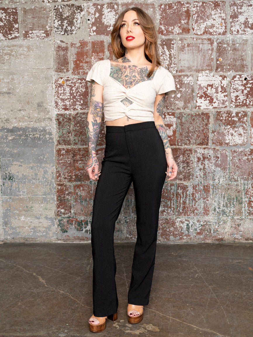 Megan Massacre wearing high-wasted black flared pants and a white top, standing against a rustic background.