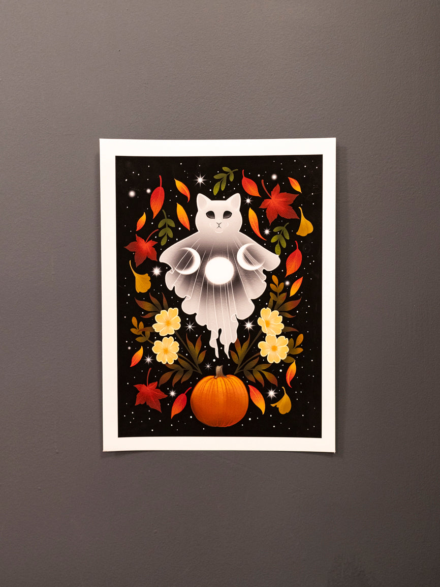 Autumn Cat Ghost art print hanging on a dark wall, displaying a white cat ghost with moons, autumn leaves, a pumpkin and florals on a black starry background. 