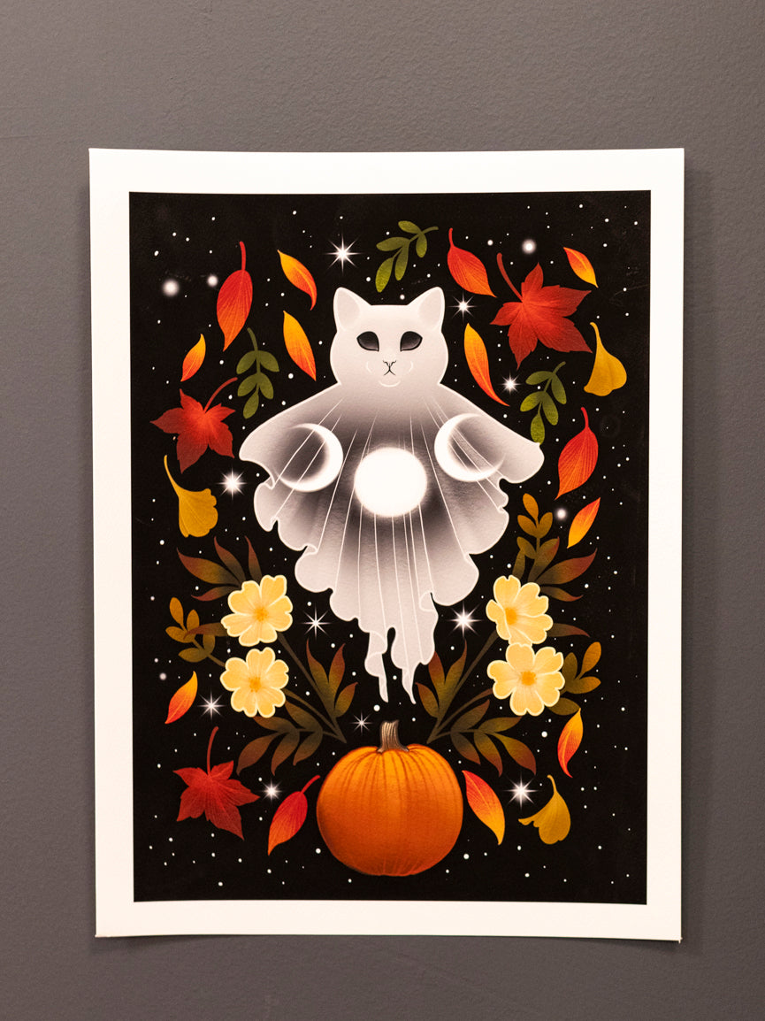 Autumn Cat Ghost art print hanging on a dark wall above an orange sofa, displaying a white cat surrounded by fall-themed decorations.