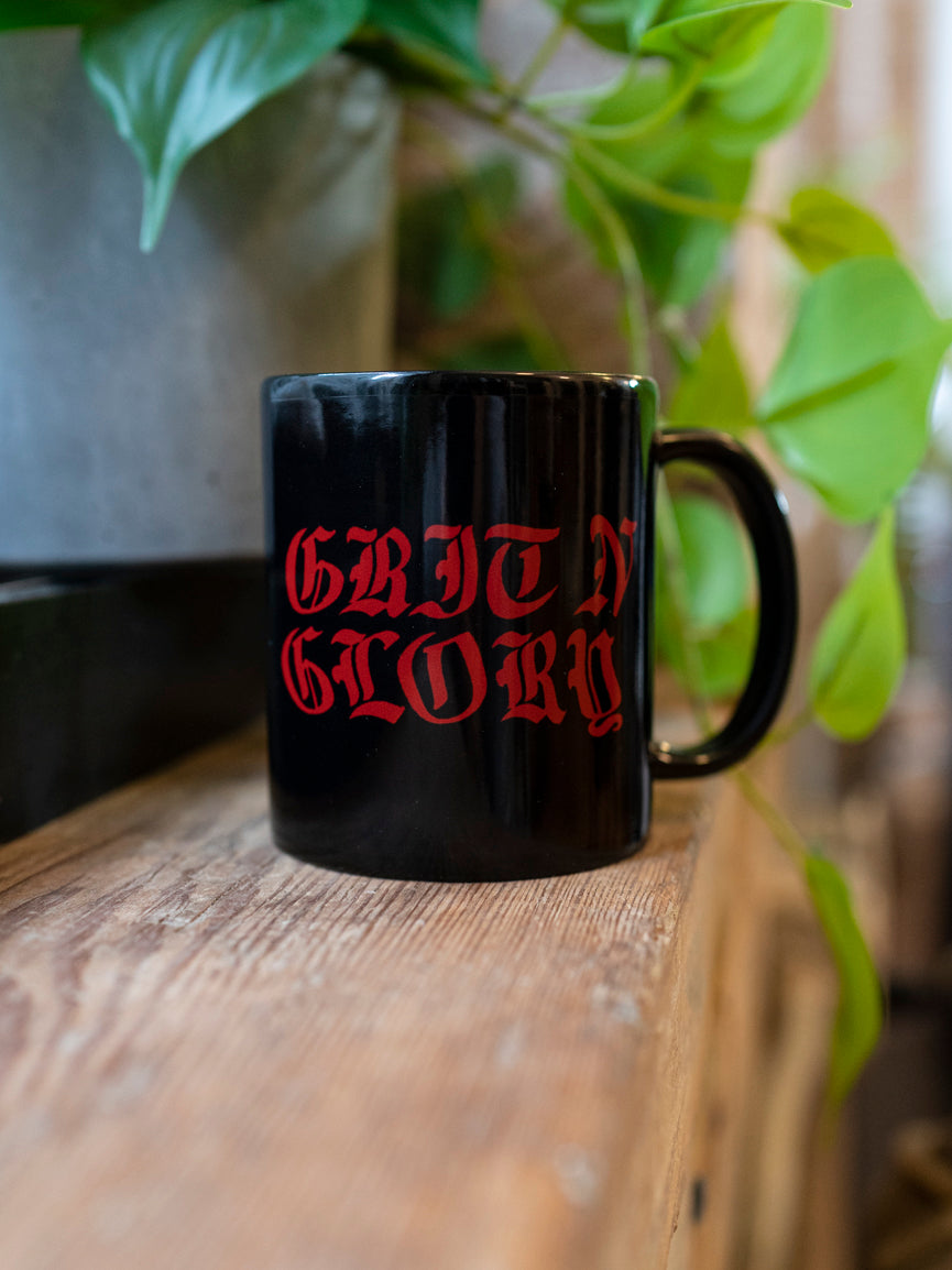 A black ceramic mug with the text 'GRIT N GLORY' written in red. The mug is placed on a wooden surface with green leaves in the background.