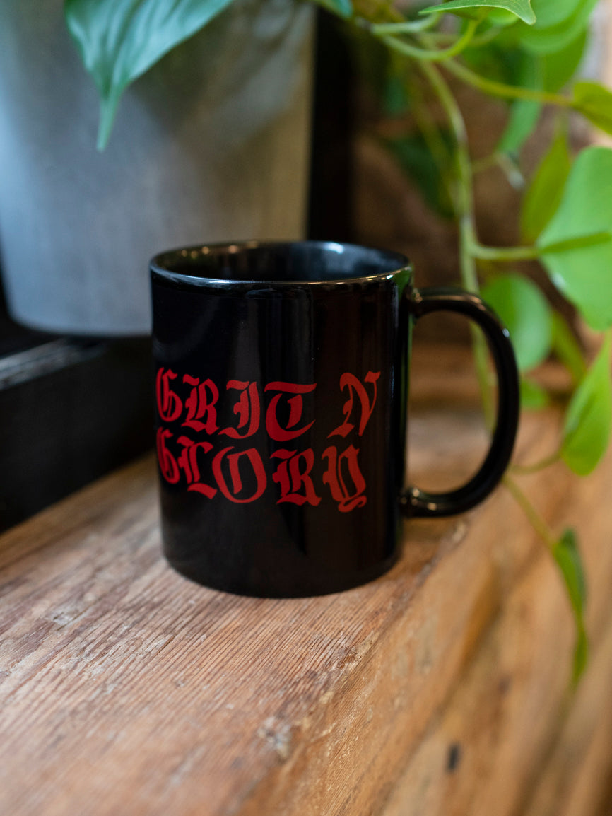 A black ceramic mug with the text 'GRIT N GLORY' written in red. The mug is placed on a wooden surface with green leaves in the background.