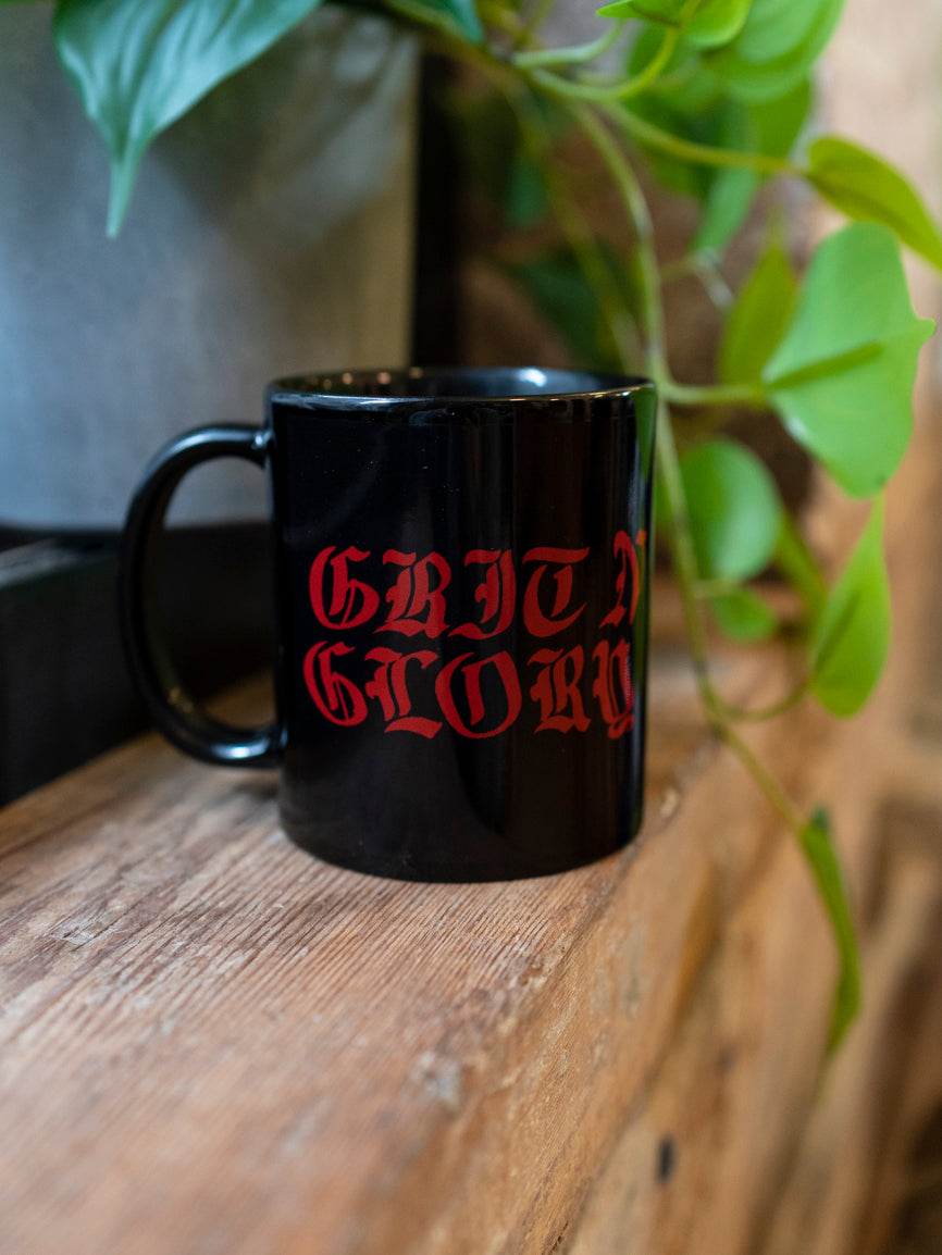 A black ceramic mug with the text 'GRIT N GLORY' written in red. The mug is placed on a wooden surface with green leaves in the background.