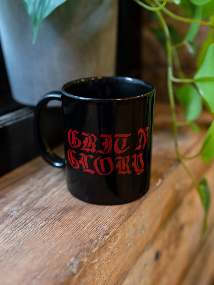 A black ceramic mug with the text 'GRIT N GLORY' written in red. The mug is placed on a wooden surface with green leaves in the background.