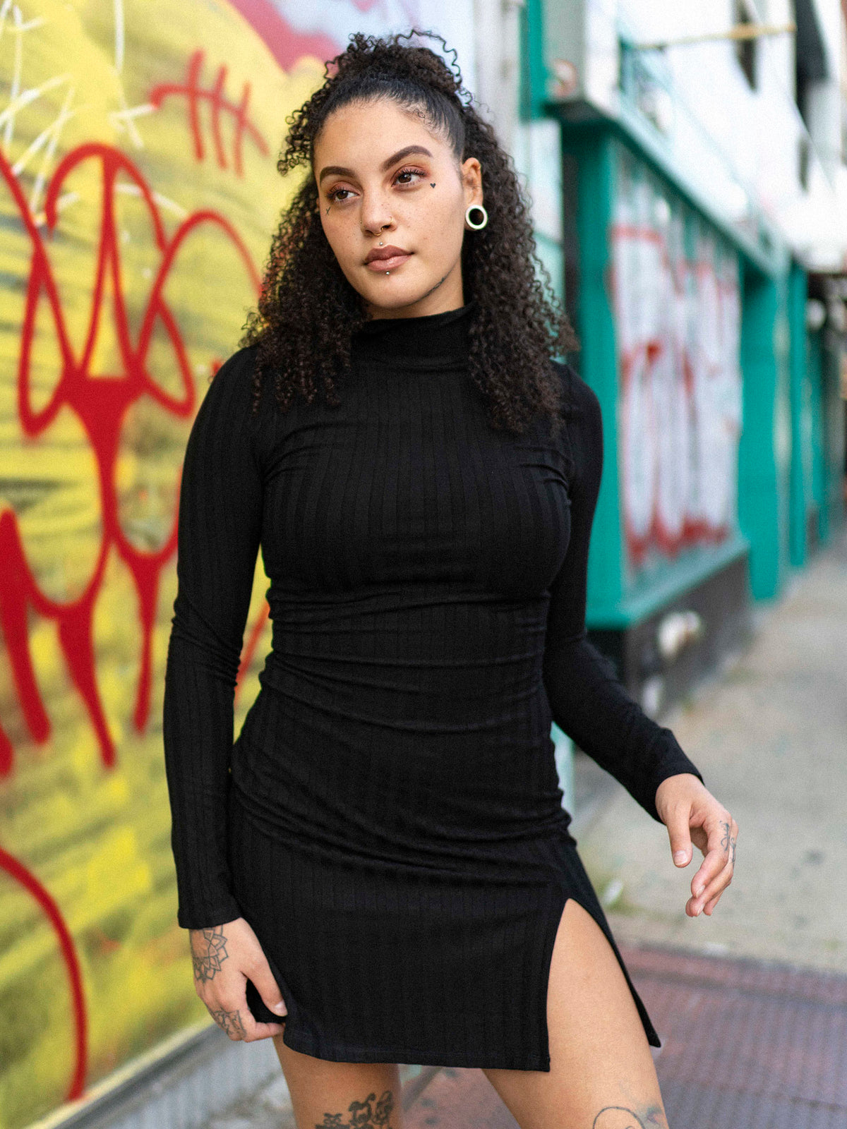A person wearing a black mini dress with a mock neck, long sleeves, and a side slit. The dress has a bodycon fit and the person is paired with black boots, standing in front of a colorful graffiti wall.