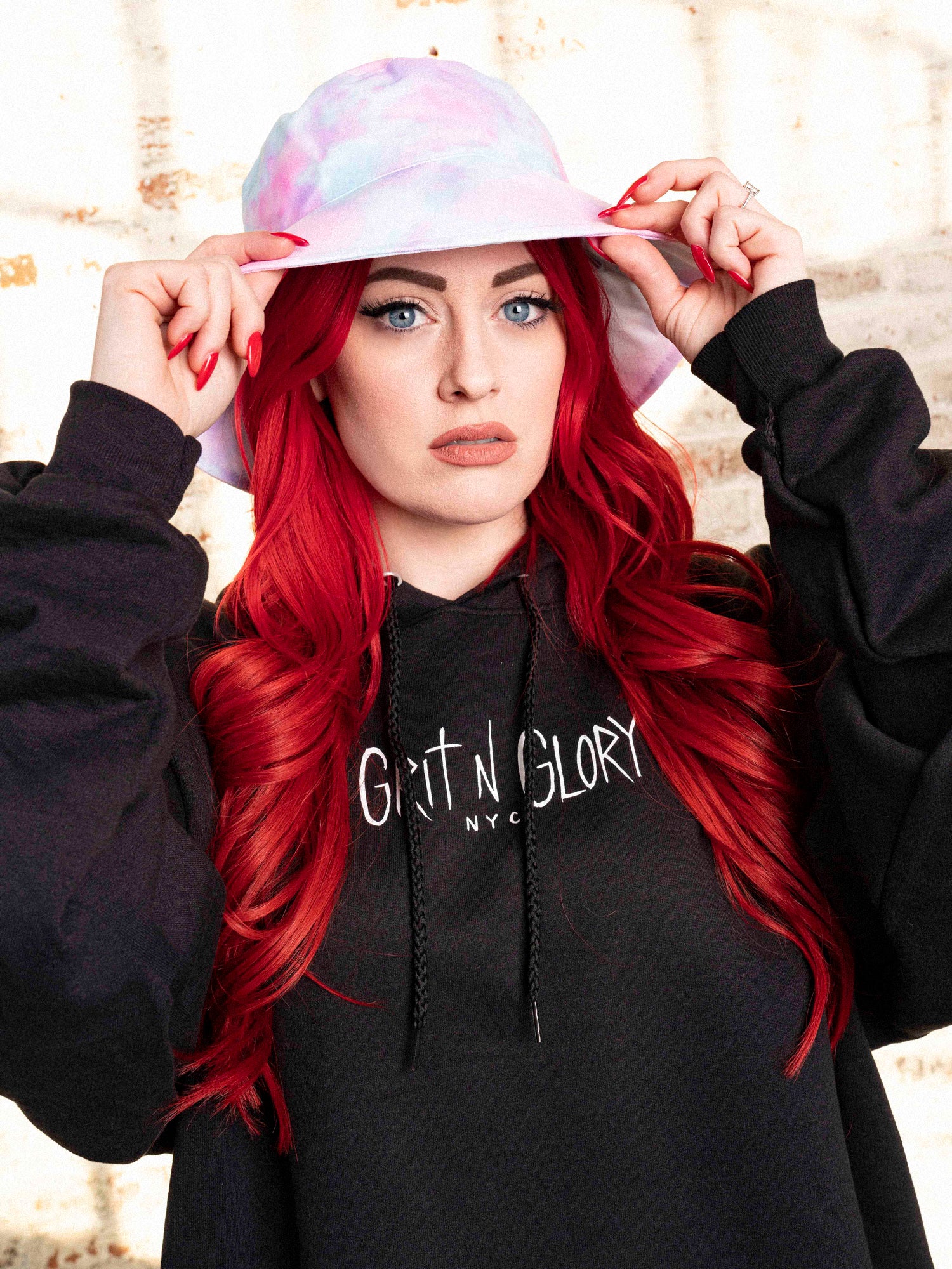 Model with bright red hair wearing a pastel tie dyed bucket hat with Grit N Glory brand black unisex hoodie. 