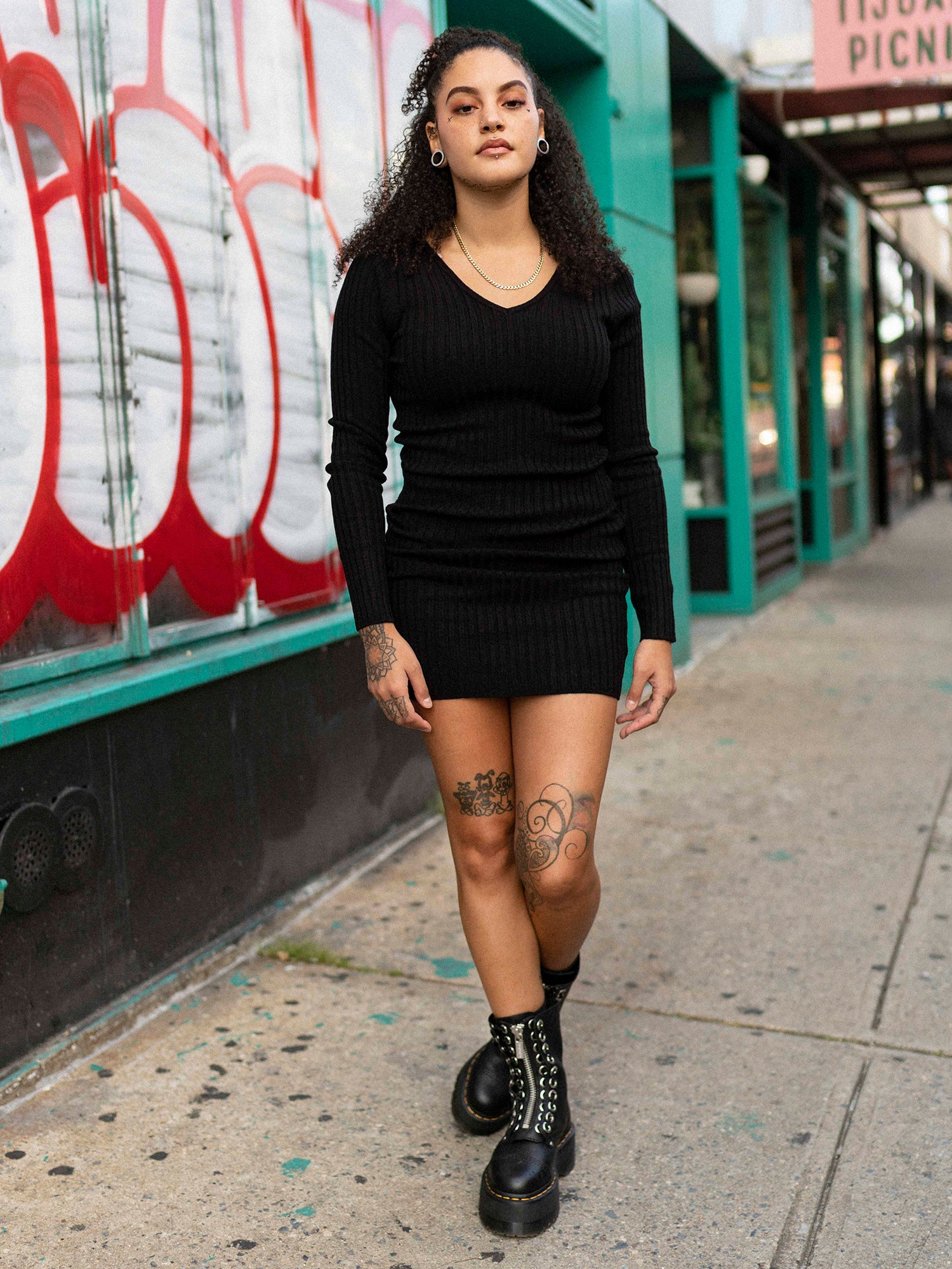 A woman standing on a sidewalk wearing a black long sleeve mini dress with a double v-neck, back neckline, and long sleeves. She is also wearing black boots and has tattoos visible on her arms.
