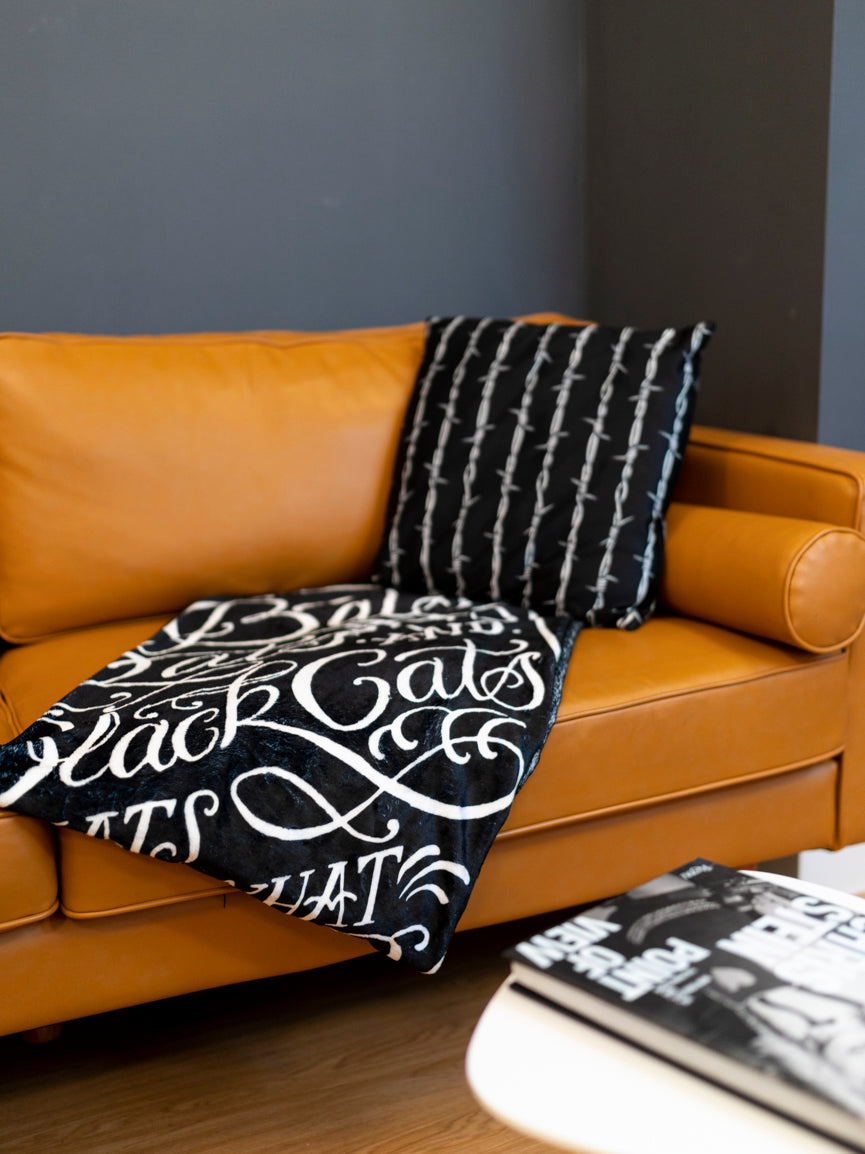 A black blanket with white text and graphic coffin design that reads 'Bats, Rats, and Black Cats - That's What Little Ghouls are Made of'. folded on a modern couch. 