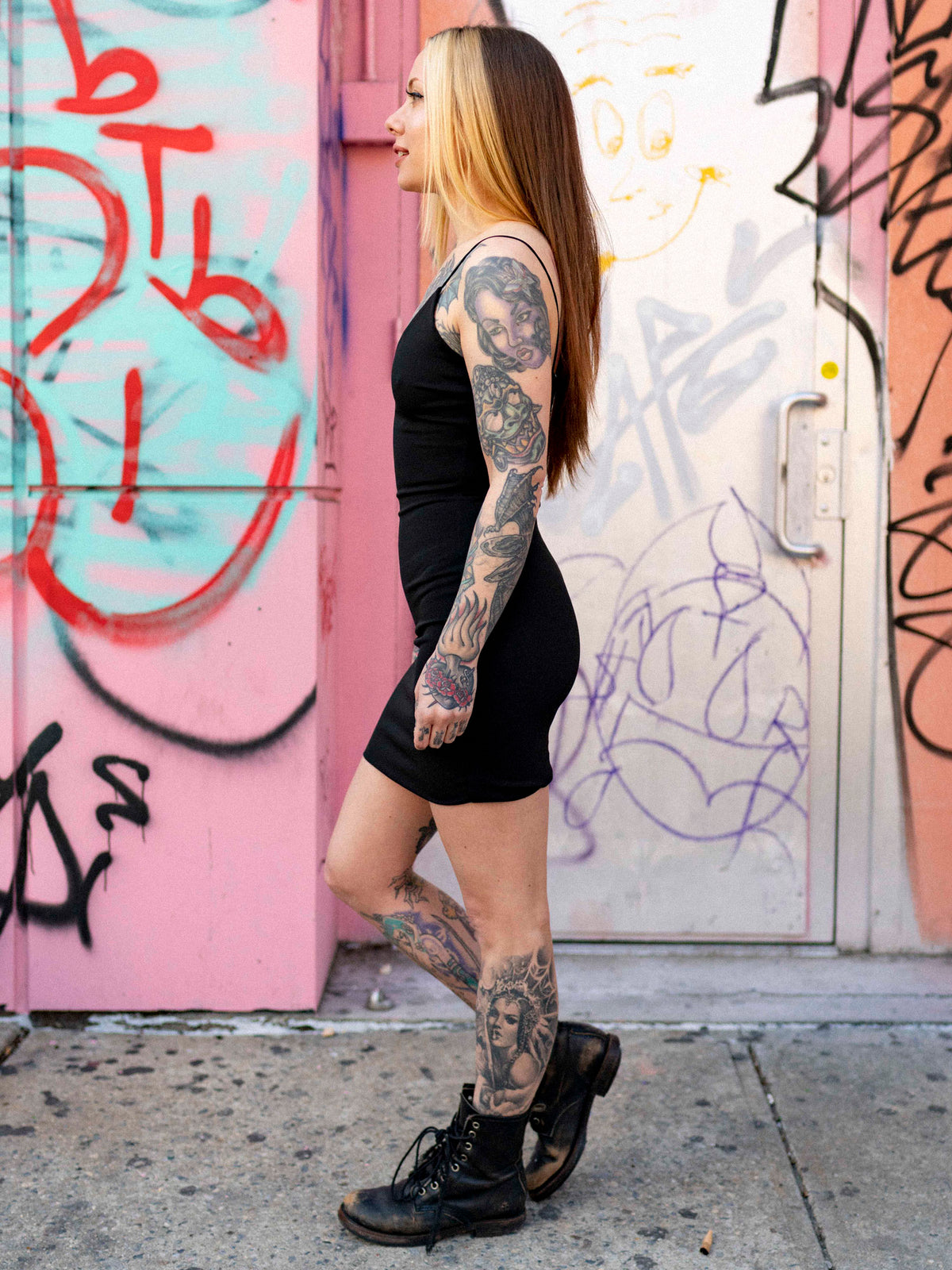 Megan Massacre standing in front of a pink wall with graffiti, wearing a black mini dress with a wide v-neck and cami straps, paired with black boots.