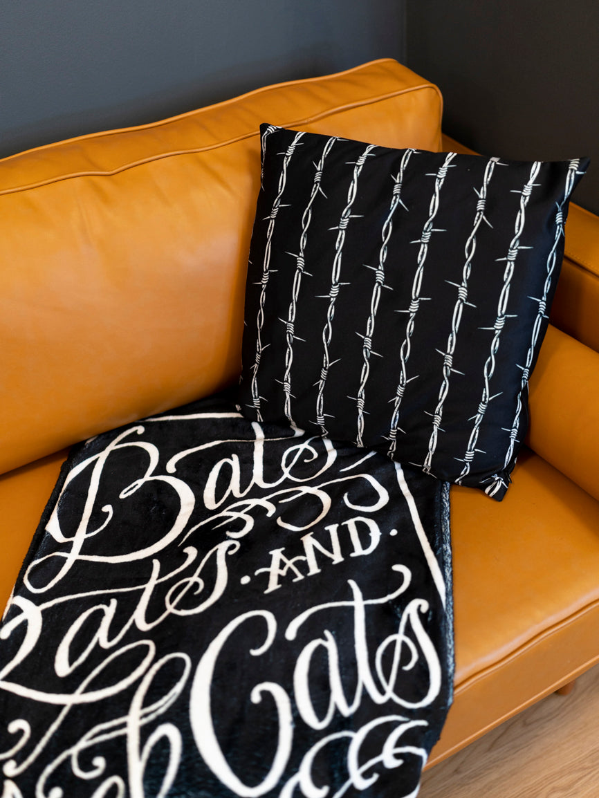 A black blanket with white text and graphic coffin design that reads 'Bats, Rats, and Black Cats - That's What Little Ghouls are Made of'. folded on a modern couch with barbed wire throw pillow. 
