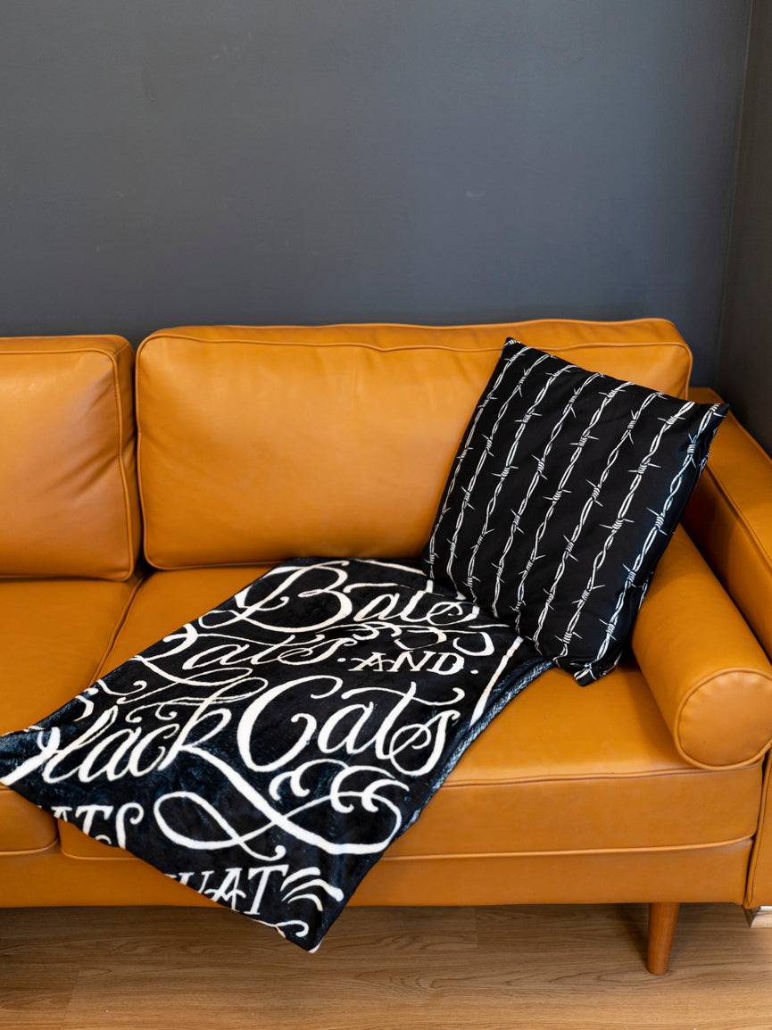 A black blanket with white text and graphic coffin design that reads 'Bats, Rats, and Black Cats - That's What Little Ghouls are Made of'. folded on a modern couch with black and white barbed wire throw pillow. 