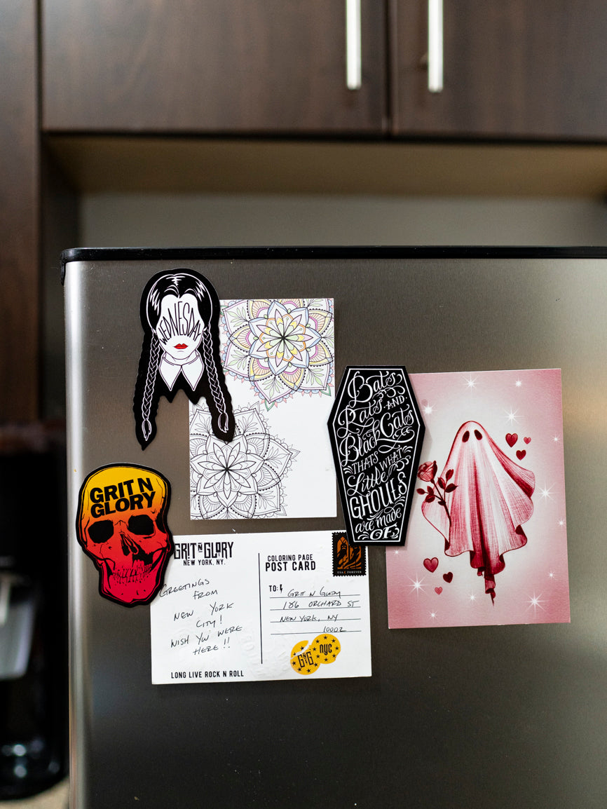 A magnet with a graphic of a Grit N Glory skull in yellow to red fade on a refrigerator with other Grit N Glory and Megan Massacre prints and magnets. 