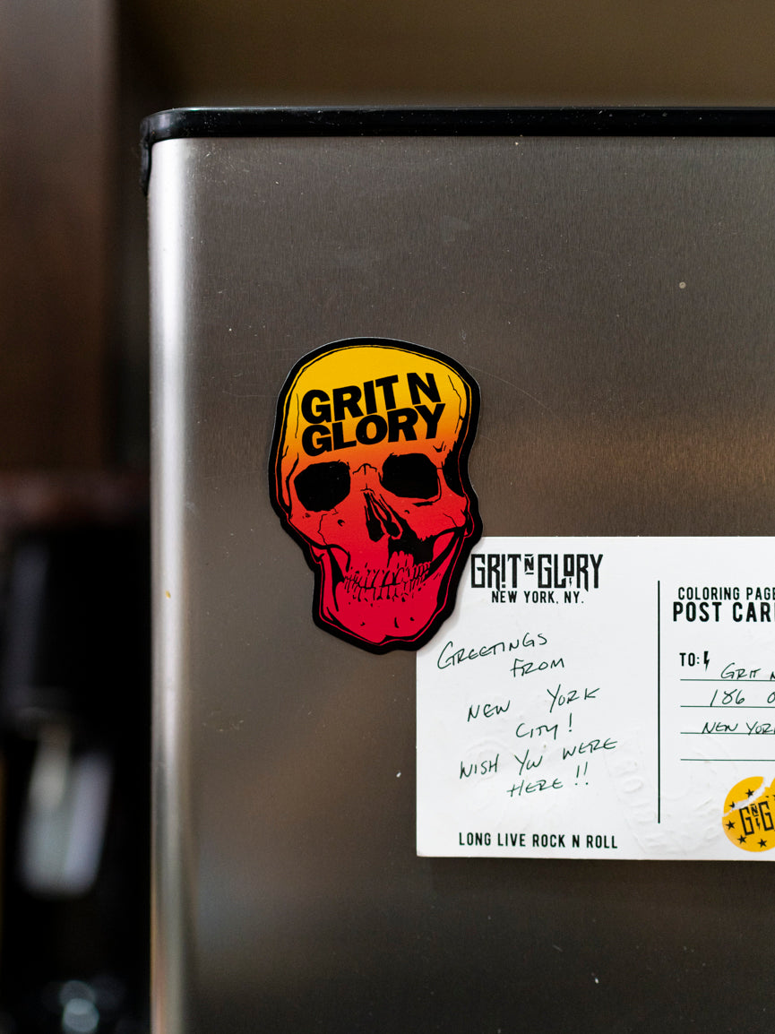 A magnet with a graphic of a Grit N Glory skull in yellow to red fade on a refrigerator.