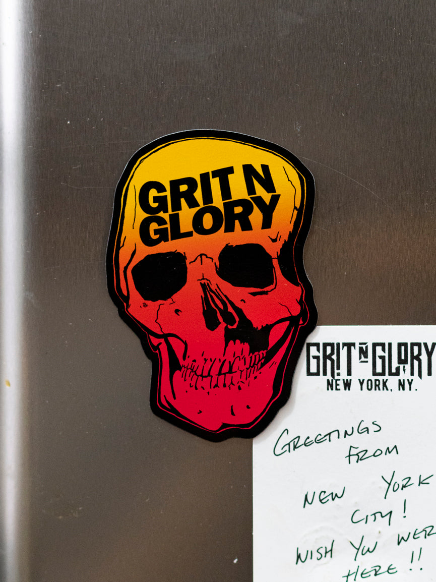 A magnet with a graphic of a Grit N Glory skull in yellow to red fade on a refrigerator.