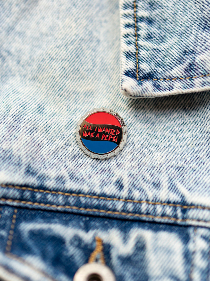 A denim jacket with a pin attached to it that has the text 'All I Wanted Was a Pepsi' on a round, red and blue background.