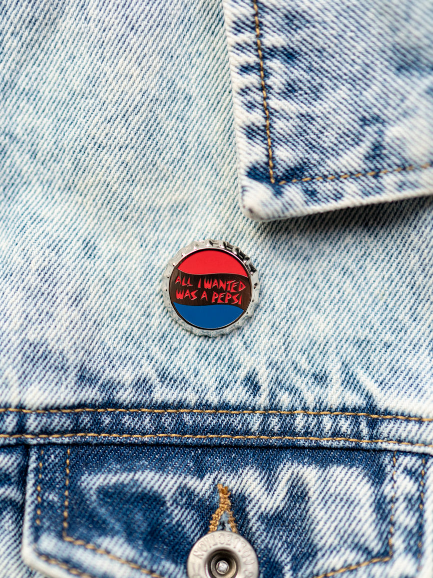 A denim jacket with a pin attached to it that has the text 'All I Wanted Was a Pepsi' on a round, red and blue background.