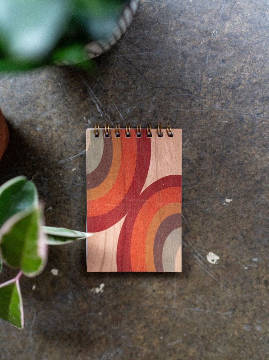 A wooden notebook with a 70's style arc pattern design on the cover, featuring a mix of orange, yellow, and brown hues, placed on a concrete surface with plants in the background.