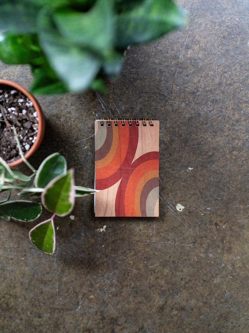 A wooden notebook with a 70's style arc pattern design on the cover, featuring a mix of orange, yellow, and brown hues, placed on a concrete surface with plants in the background.
