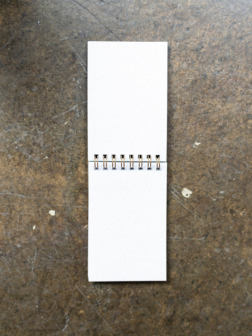 A wooden notebook with a 70's style arc pattern design on the cover, featuring a mix of orange, yellow, and brown hues, placed on a concrete surface with plants in the background.