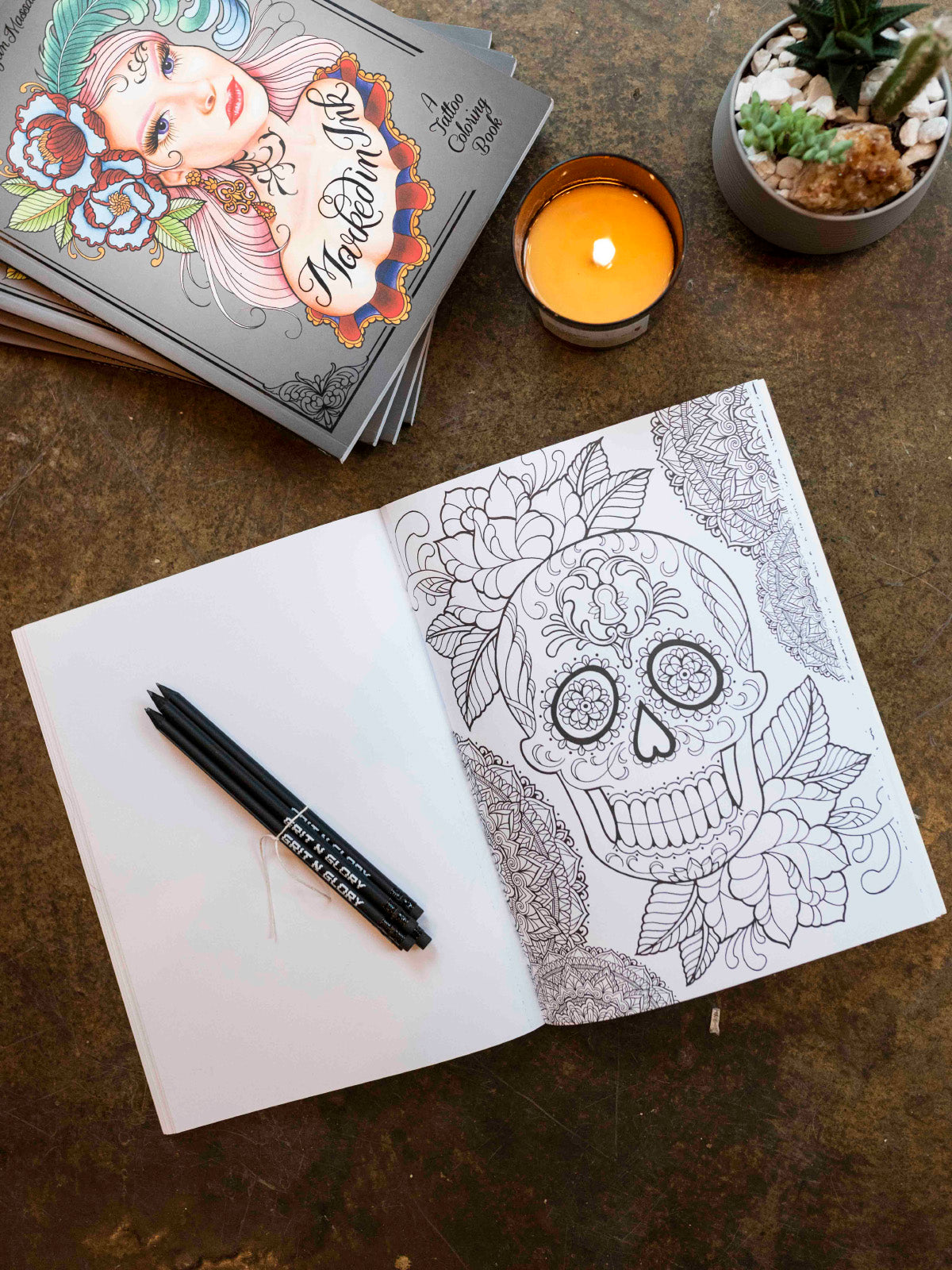 Stack of coloring books with one open featuring a coloring sheet with a sugar skull, flowers, and mandalas. 