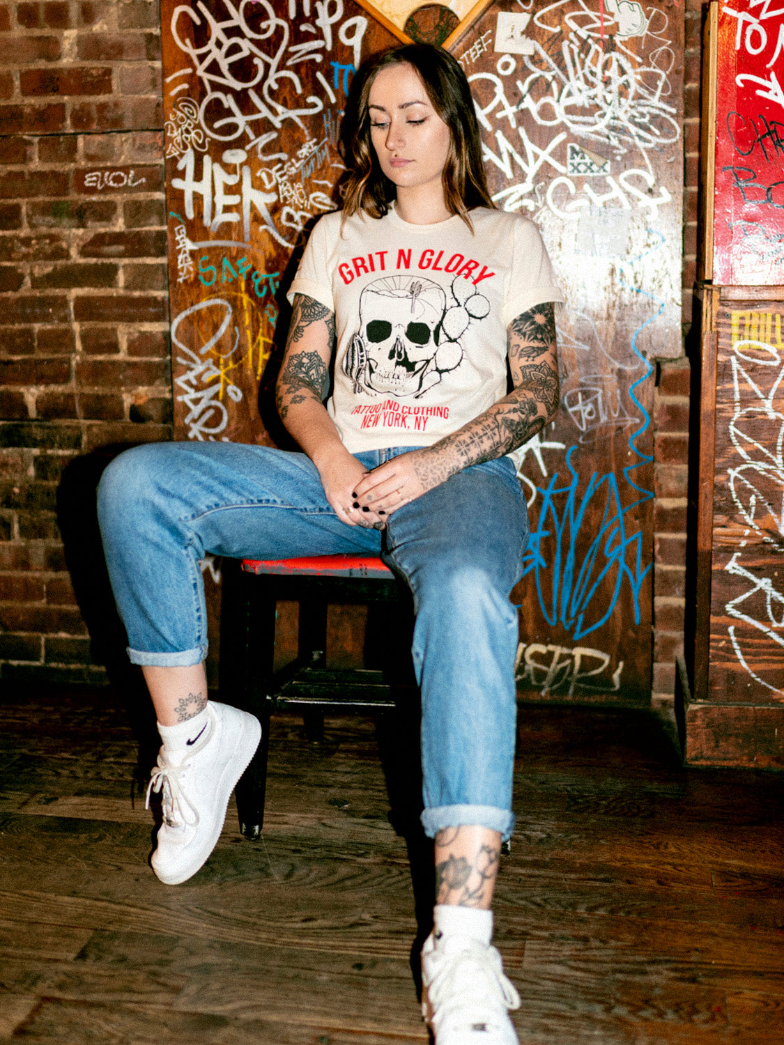 A person wearing a white crew neck t-shirt with a graphic design of a skull and text in front of a graffiti wall.