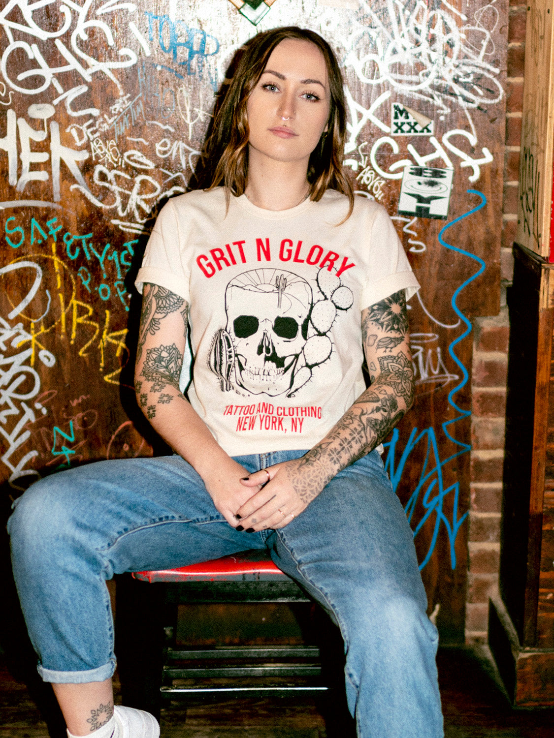 A person wearing a white crew neck t-shirt with a graphic design of a desert-themed skull and Grit N Glory red text in front of a dive bar graffiti wall.