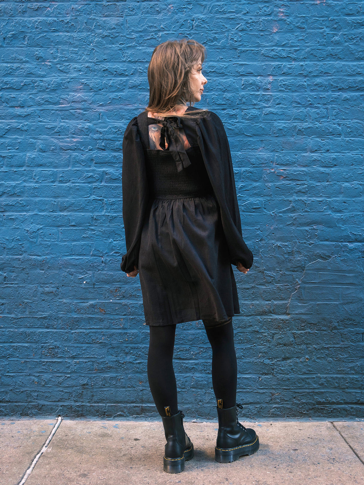 Megan Massacre wearing a black longsleeve dress against a blue brick wall
