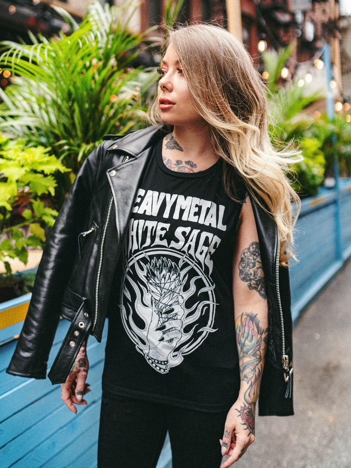 A person standing on the sidewalk wearing a black leather cropped moto jacket with a sleeveless top underneath, paired with black ripped jeans and black boots.