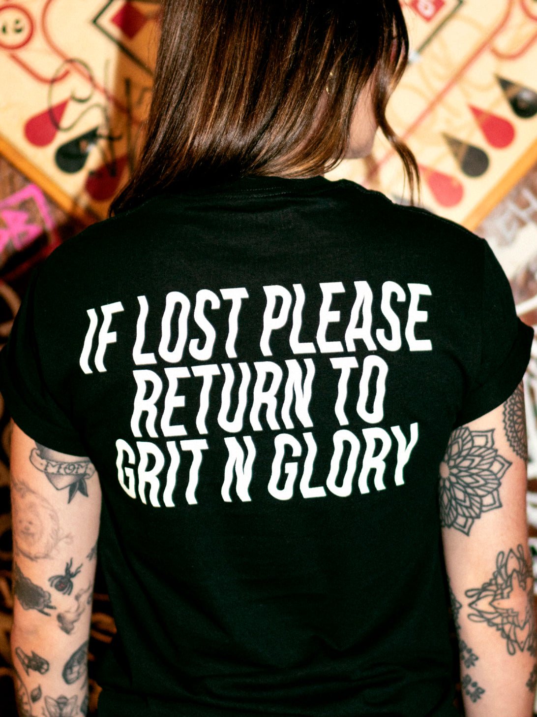 A person wearing a black graphic tee with the text 'IF LOST PLEASE RETURN TO GRIT N GLORY' on the back.