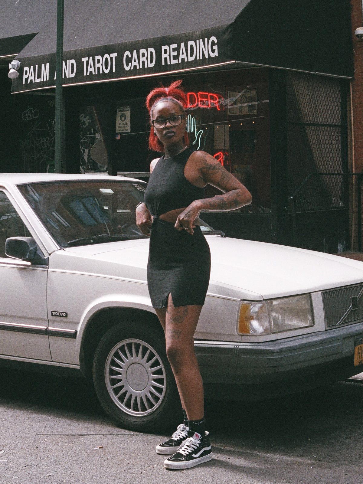 A person standing beside a car wearing a black mini dress with cutouts and a small front slit.
