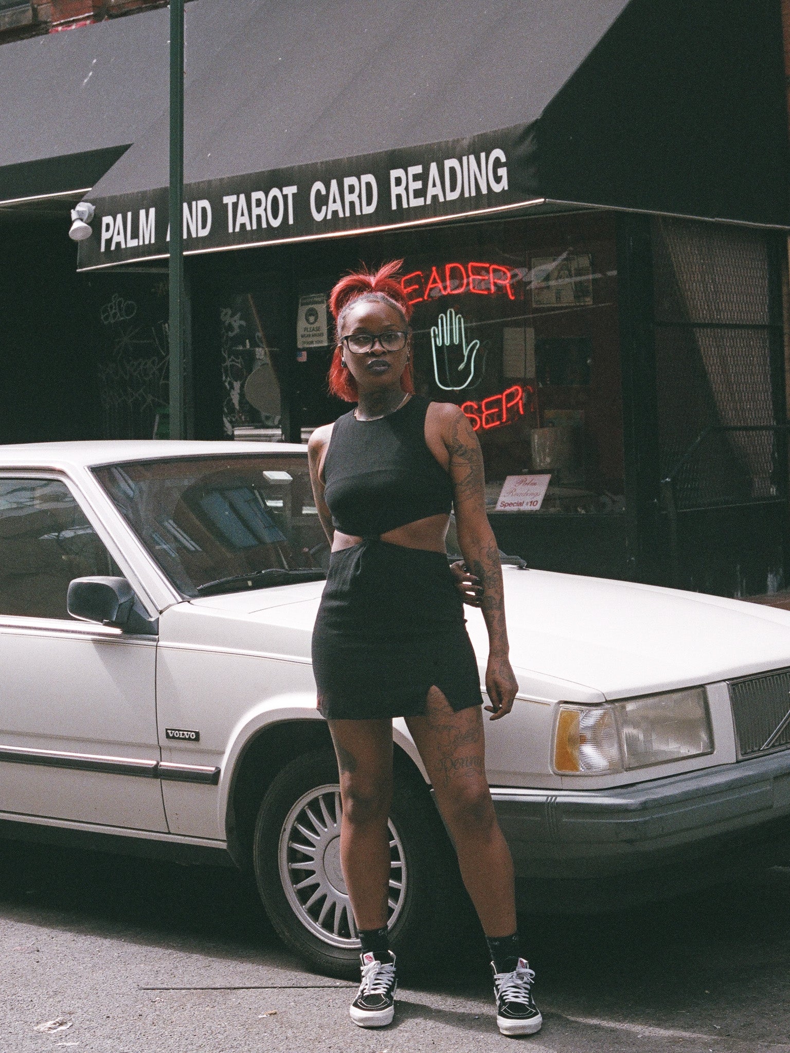 A person standing beside a car wearing a black mini dress with cutouts and a small front slit.