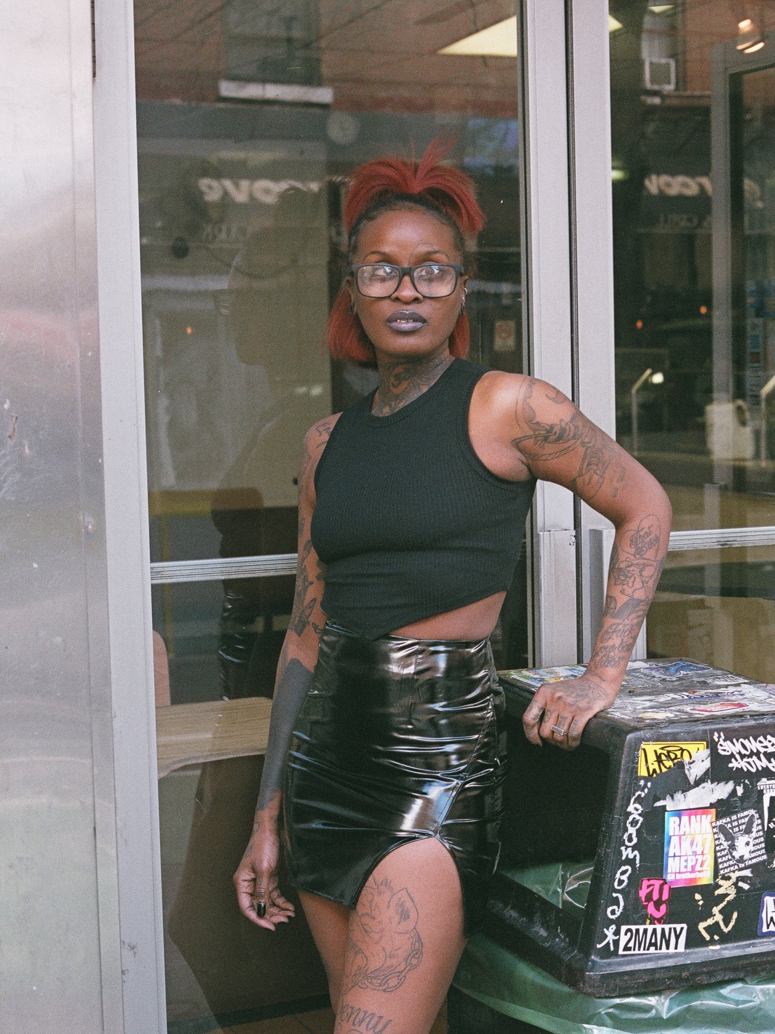 Model with red hair and glasses in a black, ribbed, tank crop top with vinyl mini skirt. 
