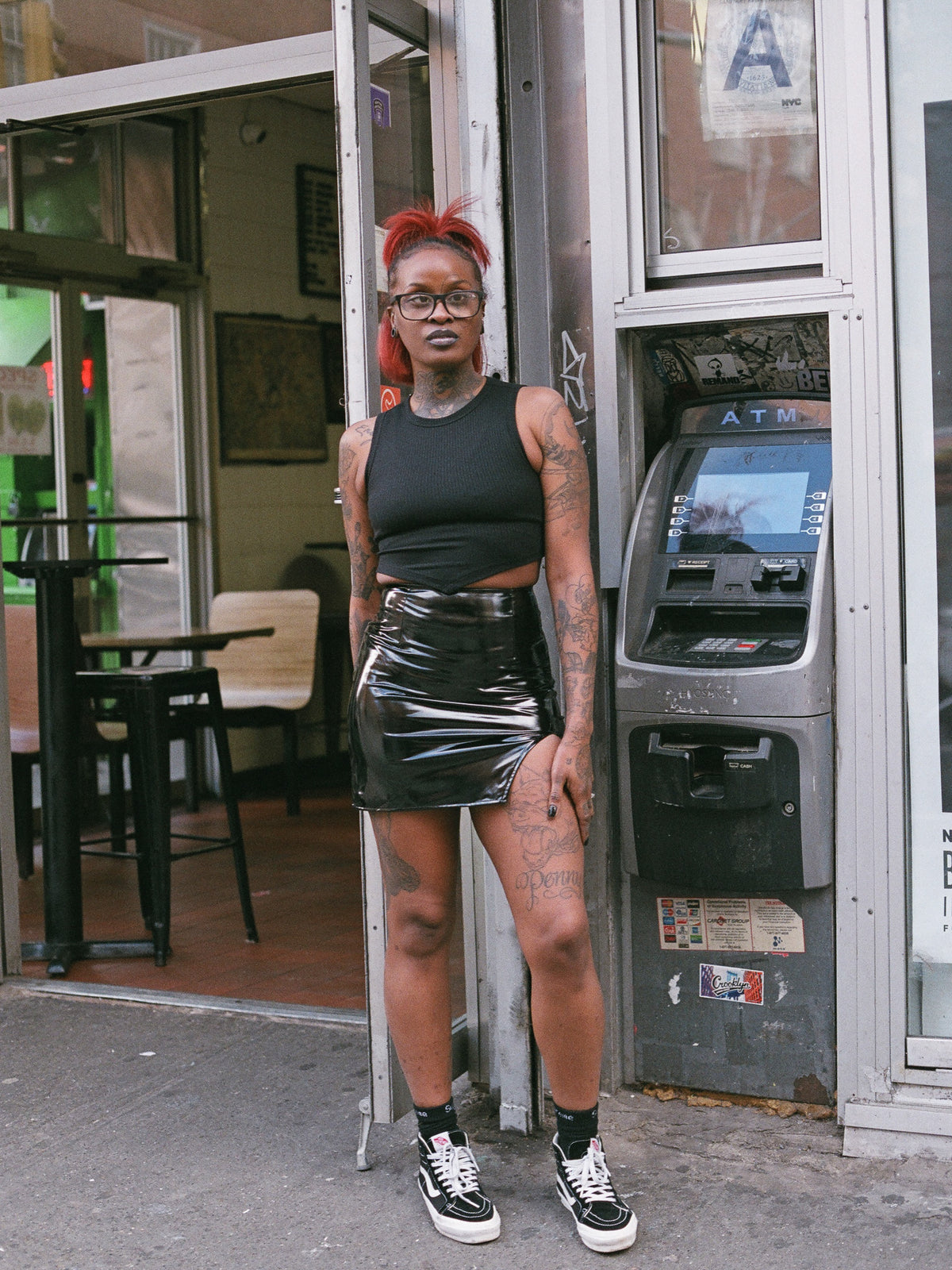 Model with red hair and glasses in a black, ribbed, tank crop top with vinyl mini skirt. 