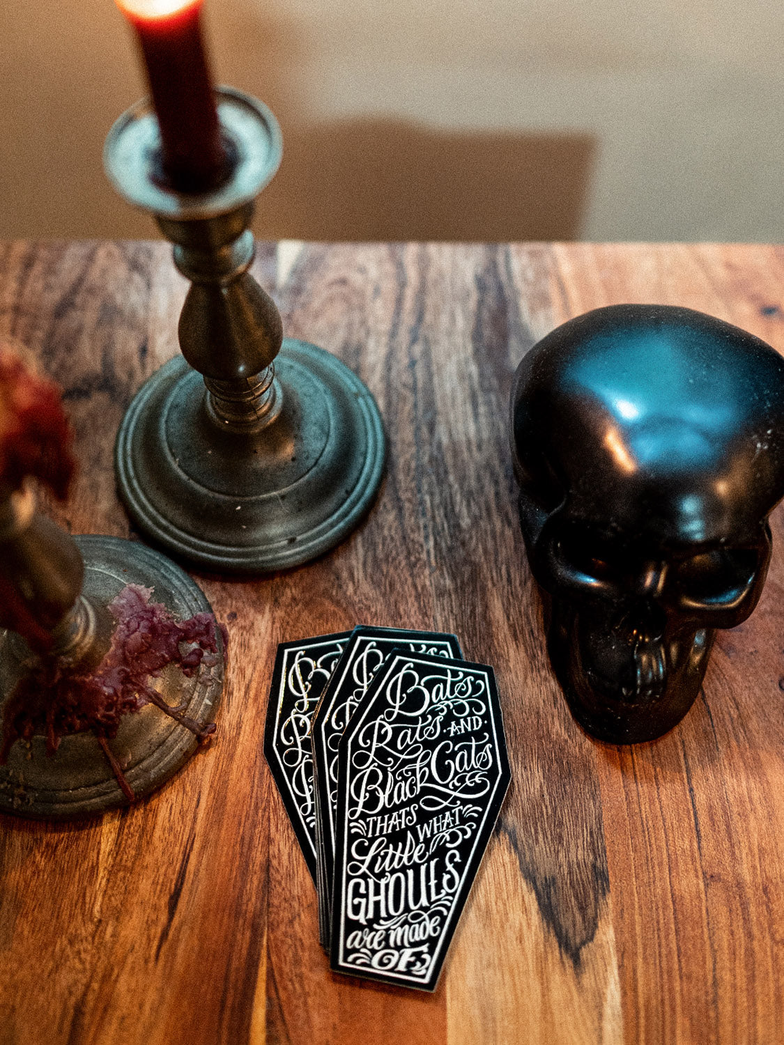 A sticker with text designed by Megan Massacre placed on a wooden surface with a skull and candle holders in the background.