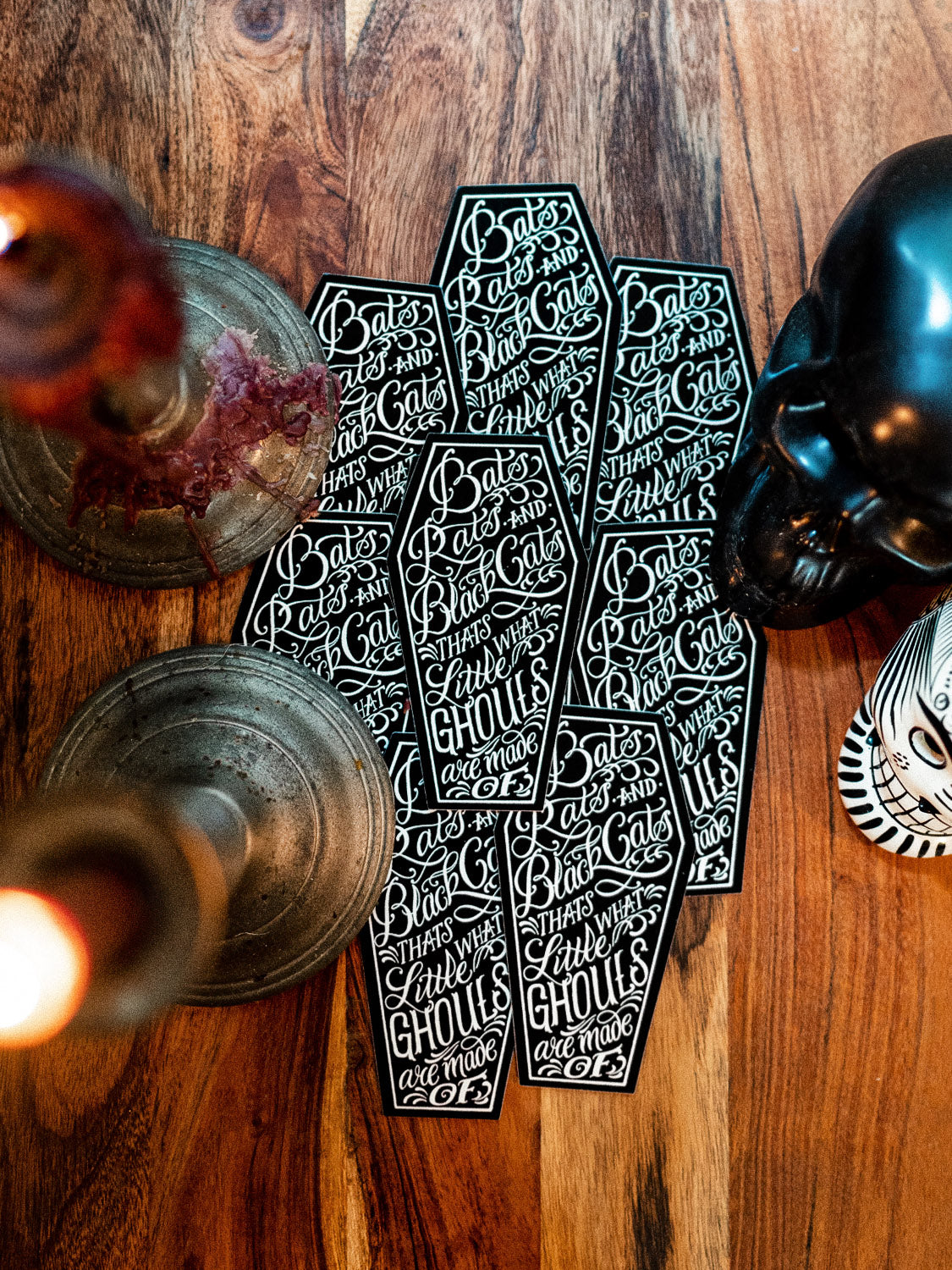A sticker with text designed by Megan Massacre placed on a wooden surface with a skull and candle holders in the background.